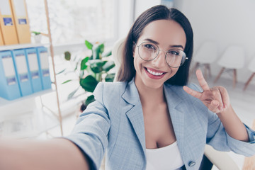 Self portrait of pretty, charming, cheerful woman in formalwear gesture v-sign to the front camera, sitting at her desk in workplace