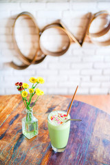 Green matcha boba or bobble tea on a wooden table with yellow flowers in a transparent vase. On the back of a blurry background, a white brick with an word - love. Bali restaurant.Side view copy space