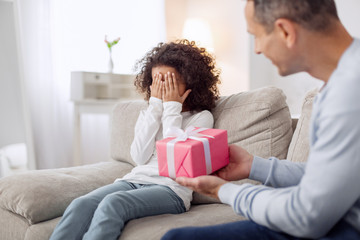 My surprise. Happy lovely curly-haired girl sitting on the sofa and closing her and her daddy holding a gift