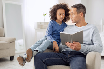 Reading together. Handsome content dark-haired father talking with his daughter and holding a book and the girl sitting near him