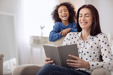 Laughter. Beautiful exuberant dark-haired young woman laughing and holding a book and her daughter standing behind her