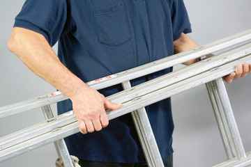 An electrician holds an aluminum ladder. The builder carries an aluminum stepladder on gray background. Worker of the plant with aluminum rung ladder