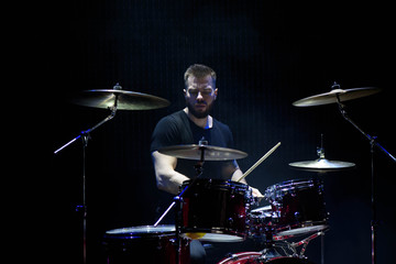 Drummer in a cap and headphones plays drums at a concert under white light in a smoke