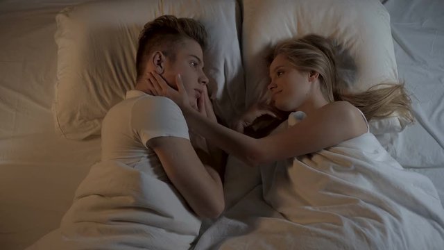 Girl looking with love at boyfriend in bed, gently stroking his face, romantic