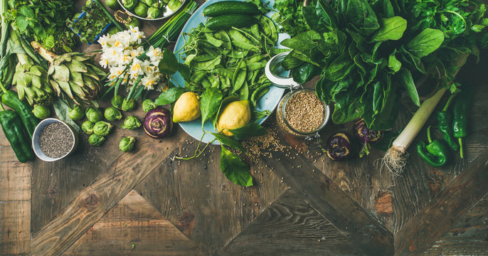 Spring healthy vegan food cooking ingredients. Flat-lay of vegetables, fruit, seeds, sprouts, flowers, greens over wooden background, top view, copy space, wide composition. Diet food concept