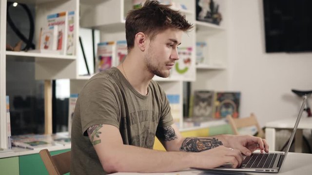 Attractive young male programmer teacher with several tattooes on his arms is mumbling something while typing on laptop keyboard.