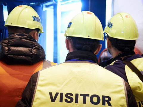 Group Of Visitors On The Factory Tour. People Go In Helmets And Uniforms For An Industrial Enterprise