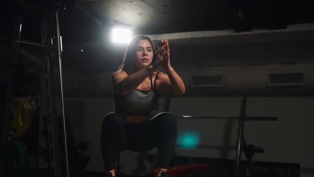 Beautiful female fitness athlete performs box jumps in a dark gym wearing black sports top and short tights with face hidden