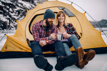 Young couple sits in tent, smiles and drinks hot tea during winter hike.