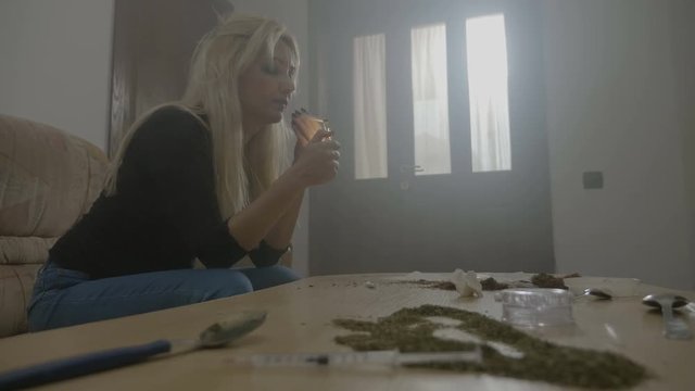 Bored attractive blonde female student sitting on the couch with table full of weed rolling herself a joint lighting and smoking it