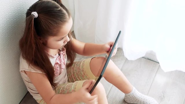 Baby little girl holding and using pc tablet and sits on the floor in room. Child learning to do makeup