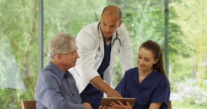 4k, A doctor and nurse having a conversation with an elderly patient in the clinic and looking at medical history on a digital touchscreen tablet.
