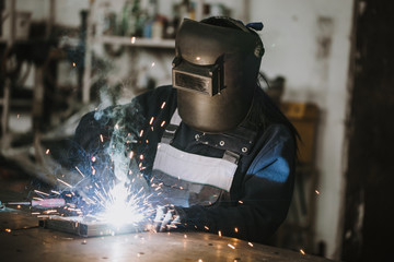Strong and worthy woman welder doing hard job in car and motorcycle repair shop. She using welding machine to fix some metal bike parts.