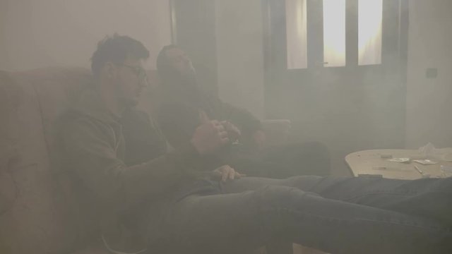 Stressed junkies male sitting in a dope house room full of smoke while inhaling and exhaling from a shared marijuana joint
