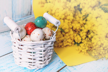 Colorful Easter eggs in a white wicker basket and Mimosa flowers on a wooden table.