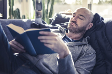 Portrait of a stylish man sitting on his sofa and reading a book after a hard day work, modern house and relaxing atmosphere. Work out situation