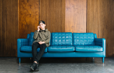Young man sitting on a blue sofa and talking on the phone. A student is sitting on the couch and...