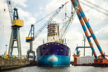 An old tanker moored in a dockyard, undergoing maintenance or repair, surrounded by large lifting...