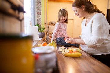 Cute charming little toddler girl sitting on the kitchen table and waiting while her pregnant mum preparing a fruit salad.