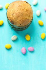 Home Baked Small Easter Cake Panettone with Raisins in Paper Form Multicolored Speckled Chocolate Candy Eggs Scattered on Blue Tabletop. Clean Minimalist Image. Top View.