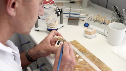 making of dental implants, male specialist in glasses with brush into arms draws white ceramics glaze on artificial jaw in workplace at table close-up
