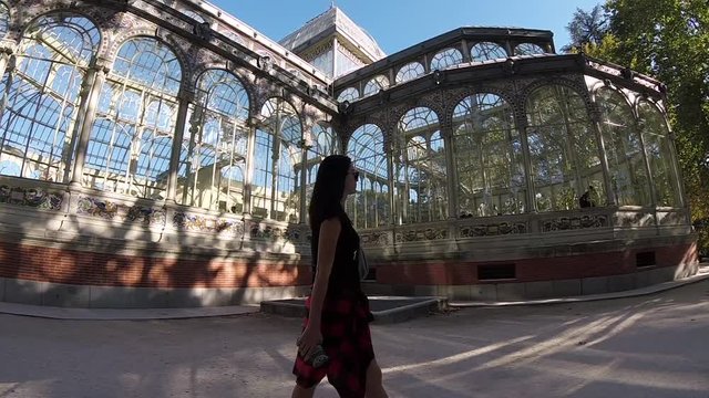 Young Woman Walking Palacio de Cristal in Madrid Buen Retiro Park. Blue Sky, Green Trees, Transparent Windows, Oriental ceramics