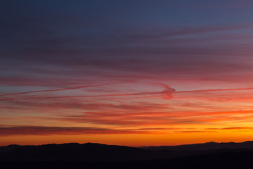 Fototapeta premium Clouds and jet vapor trails creating beautiful, colorful texture in the sky at sunset with mountain profiles on the low part of the frame