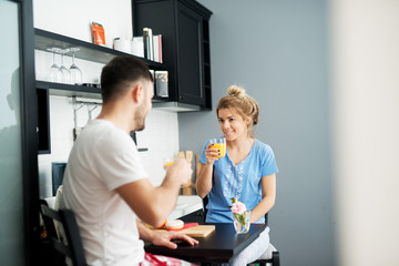 Obraz premium Young smiling couple in love having breakfast together in pajamas while drinking juice and looking flirty each other.