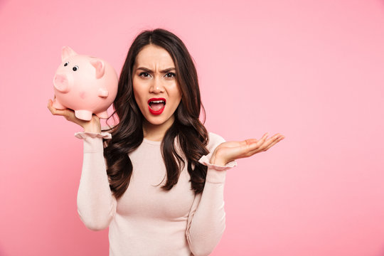 Irritated disappointed woman in casual clothing holding money box and expressing bankruptcy, isolated over pink background