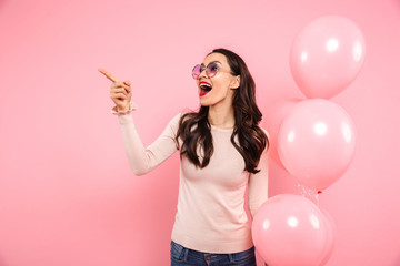 Photo of delighted adult girl with long dark hair in round glasses holding lots of balloons and pointing finger aside, isolated over pink background