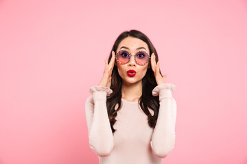 Photo of beautiful adult girl with long dark hair in round glasses expressing excitement, isolated over pink background