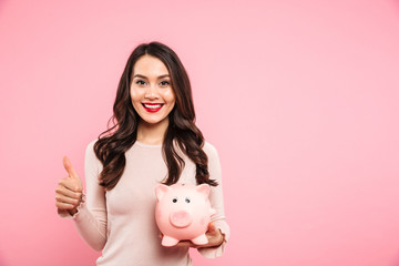 Lovely satisfied woman 20s with long dark hair holding piggybank and gesturing thumb up, isolated...