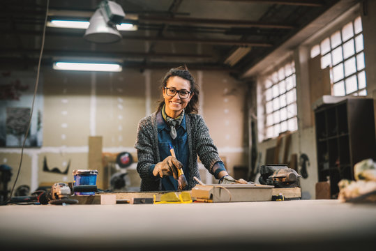 Portrait view of happy attractive hardworking middle aged professional female worker painting wood with black color in the sunny workshop or garage.