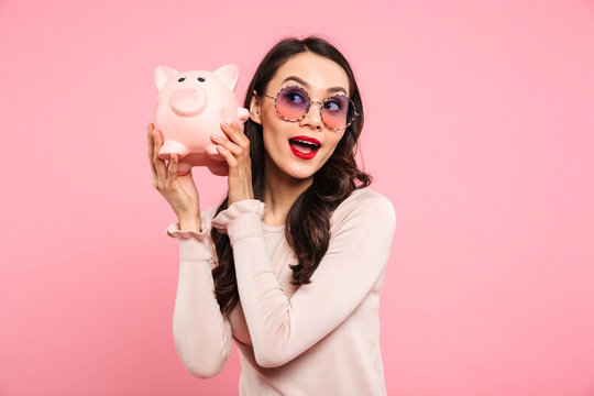 Image of lovely woman 20s with long dark hair in girlish glasses holding piggybank, isolated over pink background