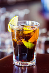 Closeup View of Glass of Cuba Libre Cocktail decorated with Lime and Lemon at lighted Bar Counter Background