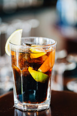 Closeup View of Glass of Cuba Libre Cocktail decorated with Lime and Lemon at lighted Bar Counter Background