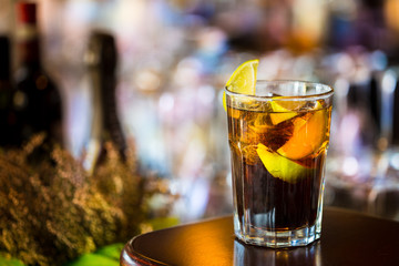 Closeup View of Glass of Cuba Libre Cocktail decorated with Lime and Lemon at lighted Bar Counter Background