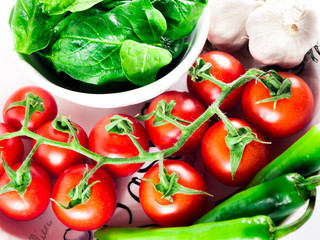 Little branch of cherry tomatoes, bowl of spinach, green hot peppers and garlic in plate over white isolated background.
