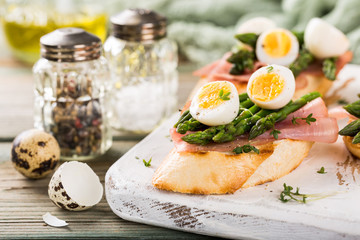 Fresh sendwich with ham, asparagus and quail eggs on white old chopping board on wooden background. Easter spring breakfast concept.