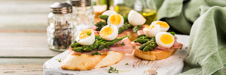 Fototapeta na wymiar Fresh sendwich with ham, asparagus and quail eggs on white old chopping board on wooden background. Easter spring breakfast concept with copy space. Banner.