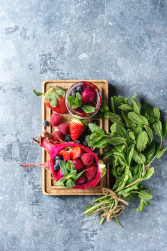 Vegan fruit salad with berries and mint served in pink dragon fruit with ingredients above on wooden serving board over blue texture background. Top view, space. Healthy eating
