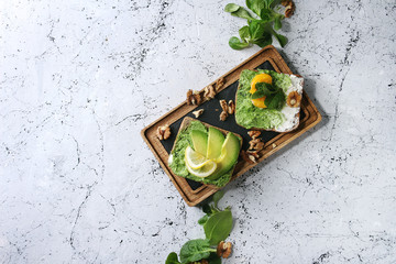 Vegetarian sandwiches with avocado, ricotta, egg yolk, spinach, walnuts on whole grain toast bread on wooden slate board with ingredients above over white marble background. Top view, copy space