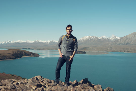Hombre joven posando en la cumbre de una montaña rodeada de agua muy azul en Nueva Zelanda