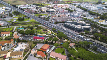 Aerial view of a wide Italian highway with three lanes in each direction. So many cars, motorcycles, trucks are running on this road near a big city and an industrial area.