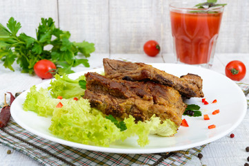 Fried pork ribs on a plate with greens and tomato juice on a white wooden background.