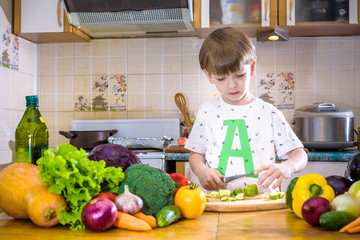 The young boy in cooking standing in the kitchen near table with vegetables