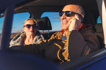 Portrait of smiling elderly couple driving car, while driver talking on phone.