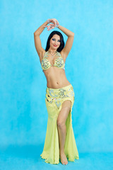 Fototapeta premium Charming dancer performs oriental belly dance on a blue background. A girl in a yellow beautiful dress is dancing an oriental belly dance on a blue background.