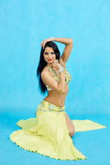 Charming dancer performs oriental belly dance on a blue background. A girl in a yellow beautiful dress is dancing an oriental belly dance on a blue background.