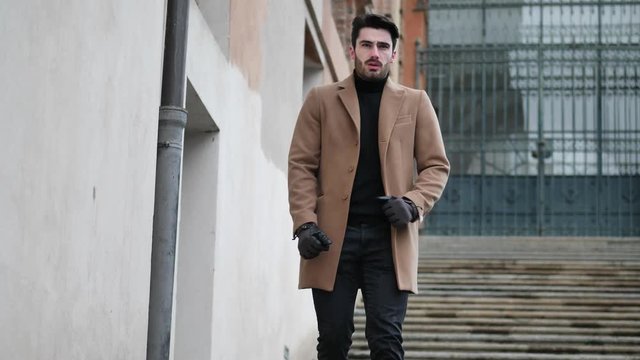 Handsome bearded young man outdoor in winter fashion, walking up marble stairs wearing black turtleneck sweater and woolen blazer jacket in city setting
